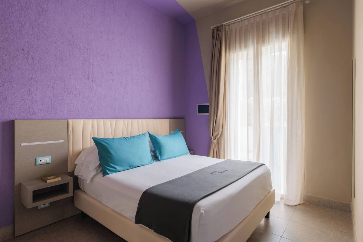 A bedroom with a bed, a nightstand, a power outlet, and a window with curtains.