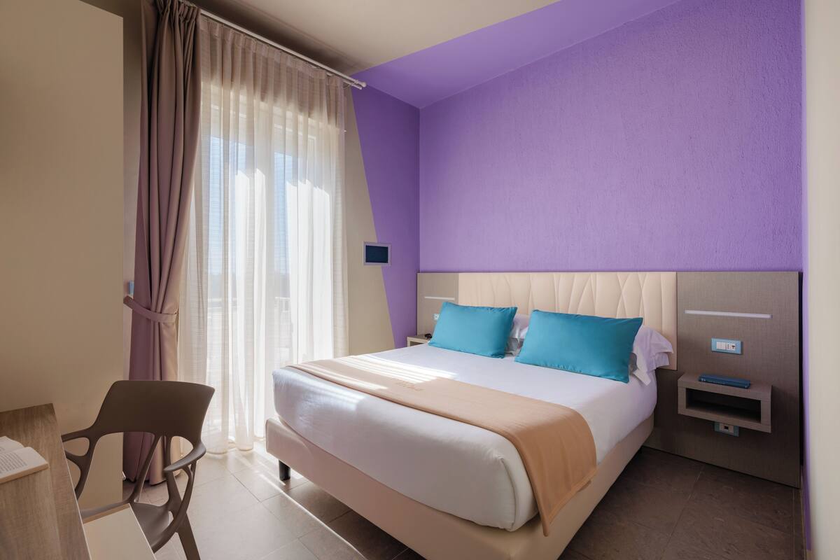 A hotel room with a bed, a desk, a chair, a nightstand, and a window with curtains.