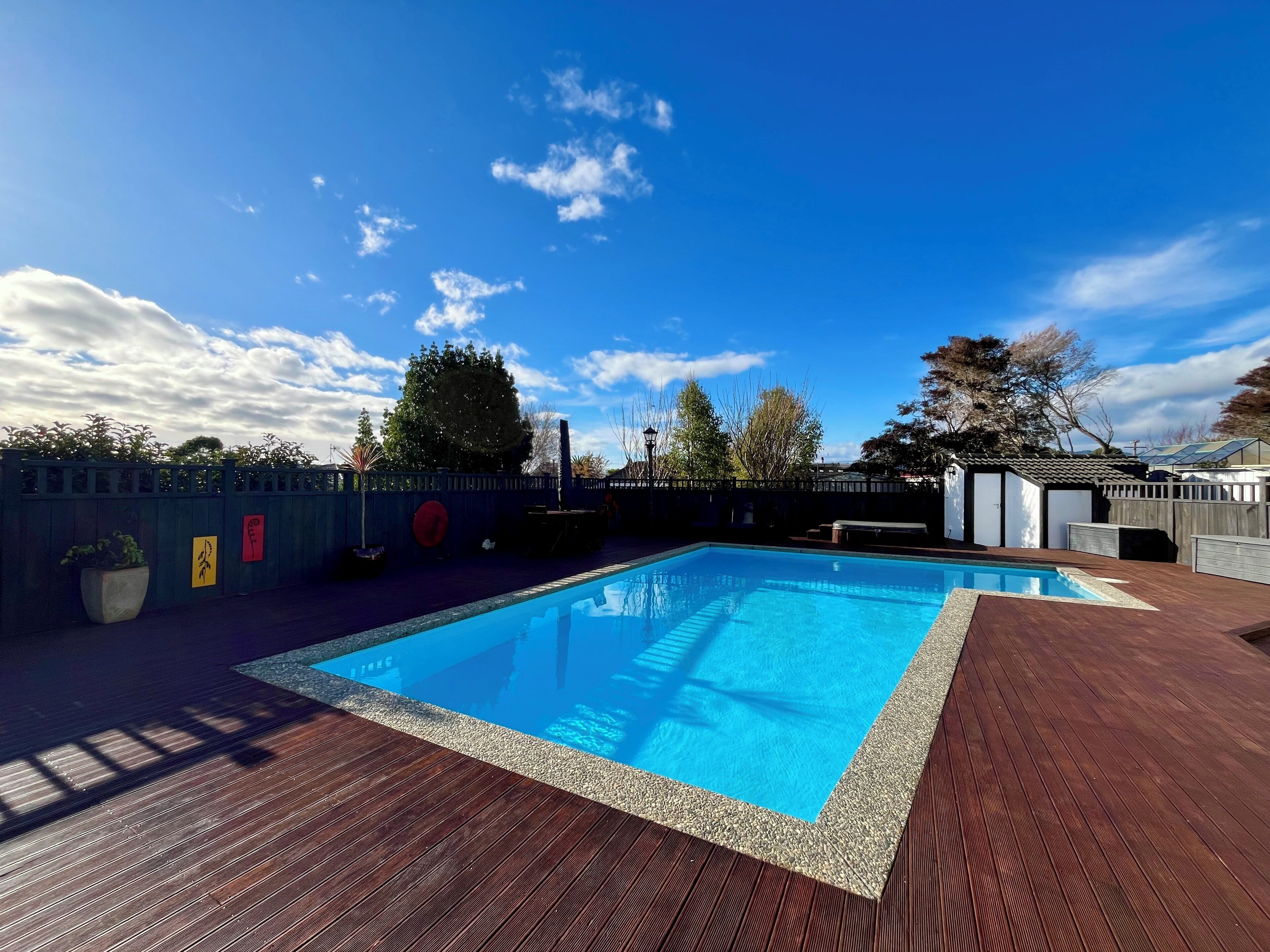 Ein rechteckiger Swimmingpool mit umlaufender Holzterrasse, einem Zaun und Bäumen im Hintergrund.