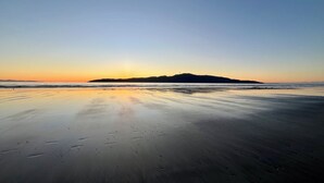 A beach with footprints leading to the water, a calm sea, and a distant mountain silhouette during sunset. - Tudor Manor Bed & Breakfast (Paraparaumu)