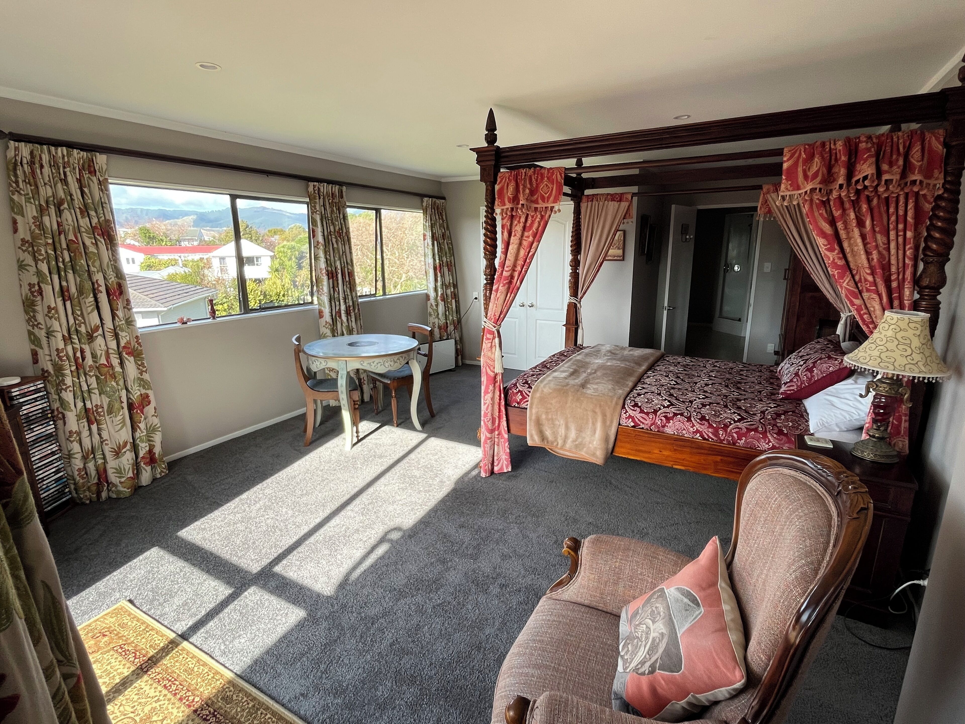 A bedroom with a four-poster bed, a sitting area with a table and chairs, and a view of the outdoors through large windows.