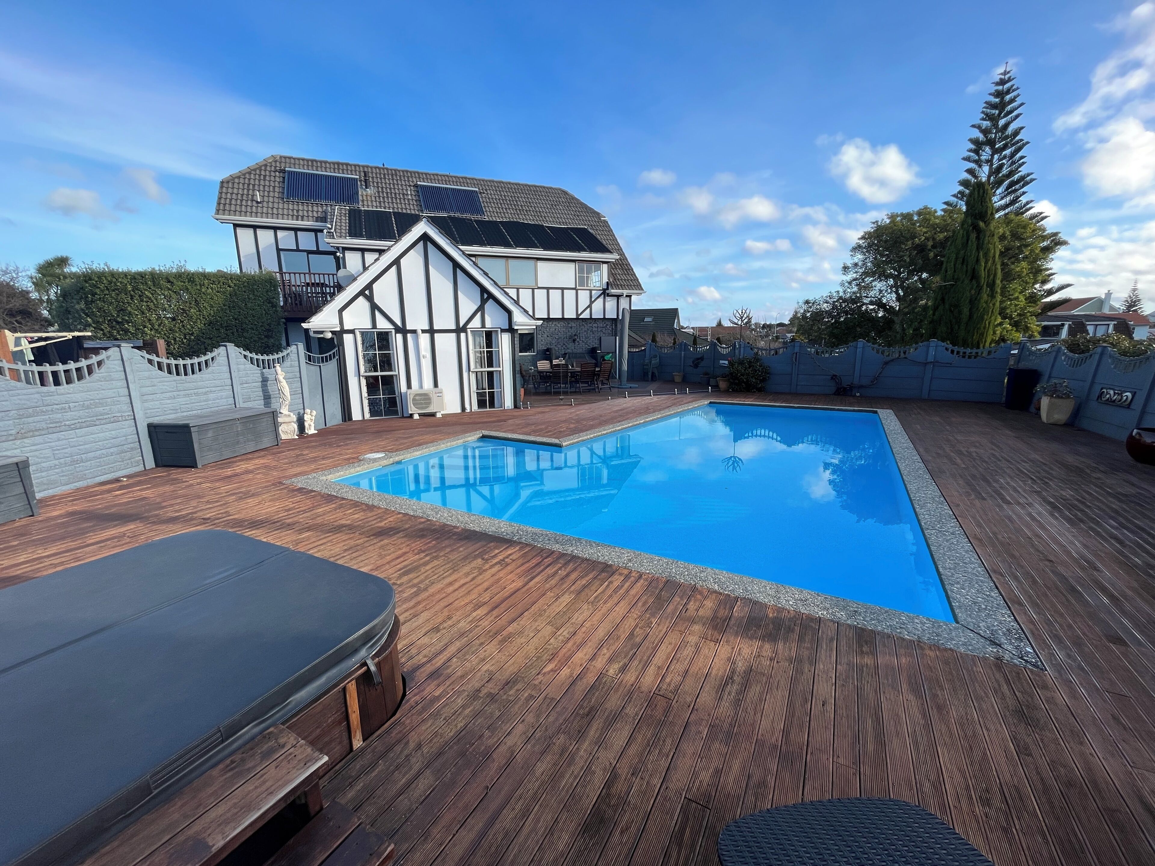 A swimming pool with a wooden deck, a lounge chair, and a building with solar panels in the background.