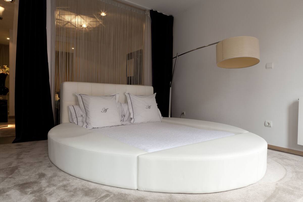 Quarto moderno com cama curva, luminária de chão grande e cortinas translúcidas.