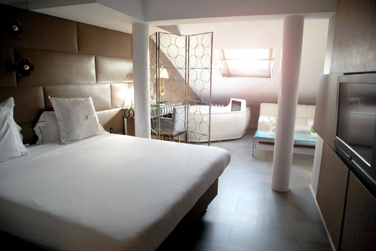 Quarto de hotel moderno com uma cama grande, área de estar com sofá e cadeiras, divisória de vidro e uma televisão.