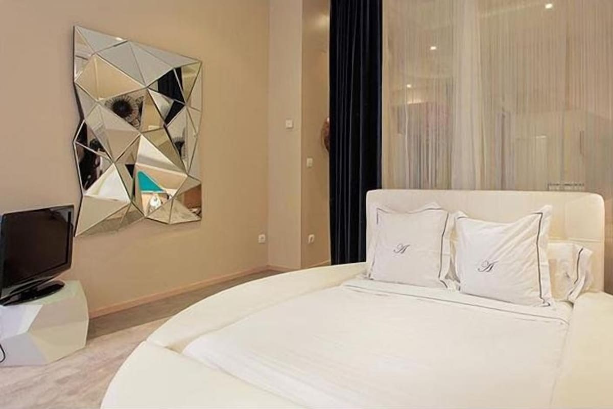 A modern hotel room with a large bed, a geometric mirror, a television, and a lamp.