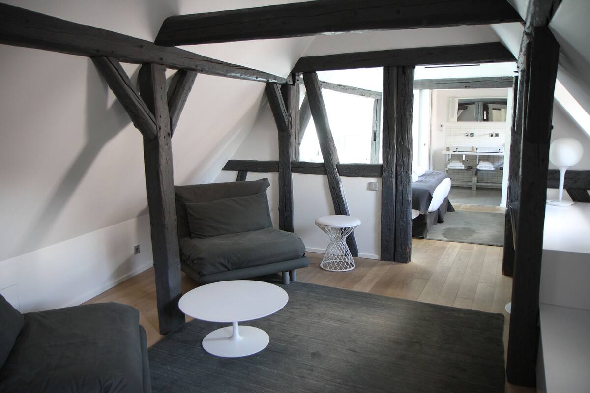 A loft-style room with exposed wooden beams, a gray sofa, a small round table, and a view into the kitchen area.