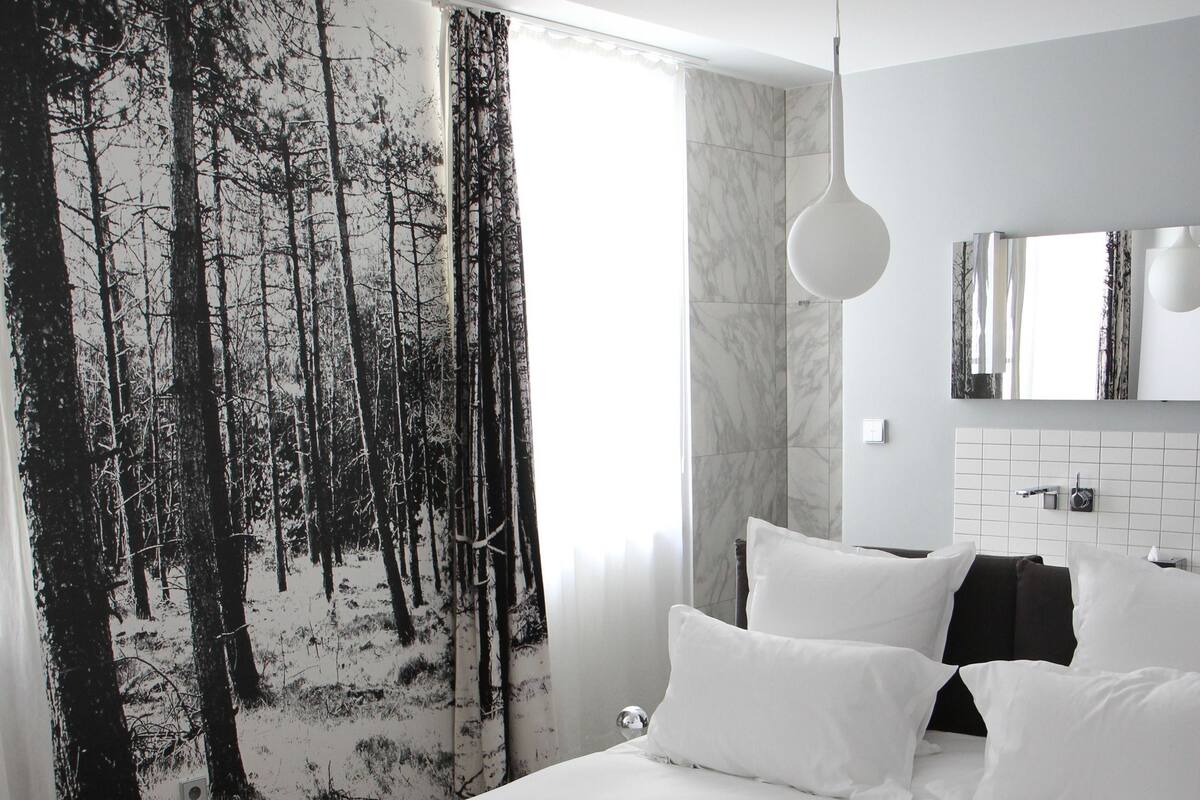 A hotel room with a bed, white pillows, a black and white blanket, a forest-themed wall, a hanging light, and a bathroom with a mirror and sink.