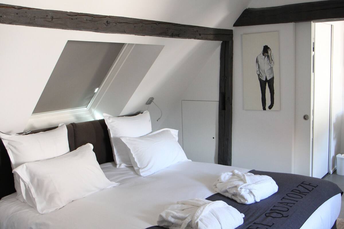 A neatly made bed with white linens and a black blanket, a skylight above, and a framed picture on the wall.