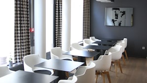 A modern dining area with white chairs and black tables, large windows with patterned curtains, and a wall-mounted artwork. - Hôtel Quatorze (Colmar)