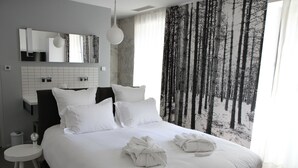 A modern hotel room with a large bed, white bedding, a round bedside table, a mirror, and a forest-themed window view. - Hôtel Quatorze (Colmar)