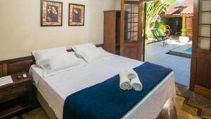 A hotel room with a bed, two framed pictures on the wall, a wooden nightstand, and a view of the outdoor area including a pool and plants. - Pousada Acquarela Paraty (Paraty)