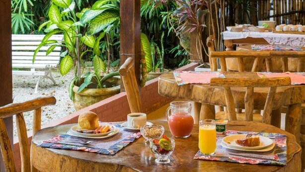 A wooden table with breakfast items including toast, fruit, and a drink, set in a tropical garden with a bench and potted plants in the background. - Pousada Acquarela Paraty (Paraty)
