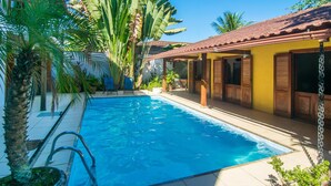 A swimming pool with a tiled deck, surrounded by tropical plants and a yellow building with wooden shutters. - Pousada Acquarela Paraty (Paraty)