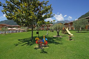 A playground with a swing set and a slide, surrounded by well-maintained grass and trees, with buildings in the background.