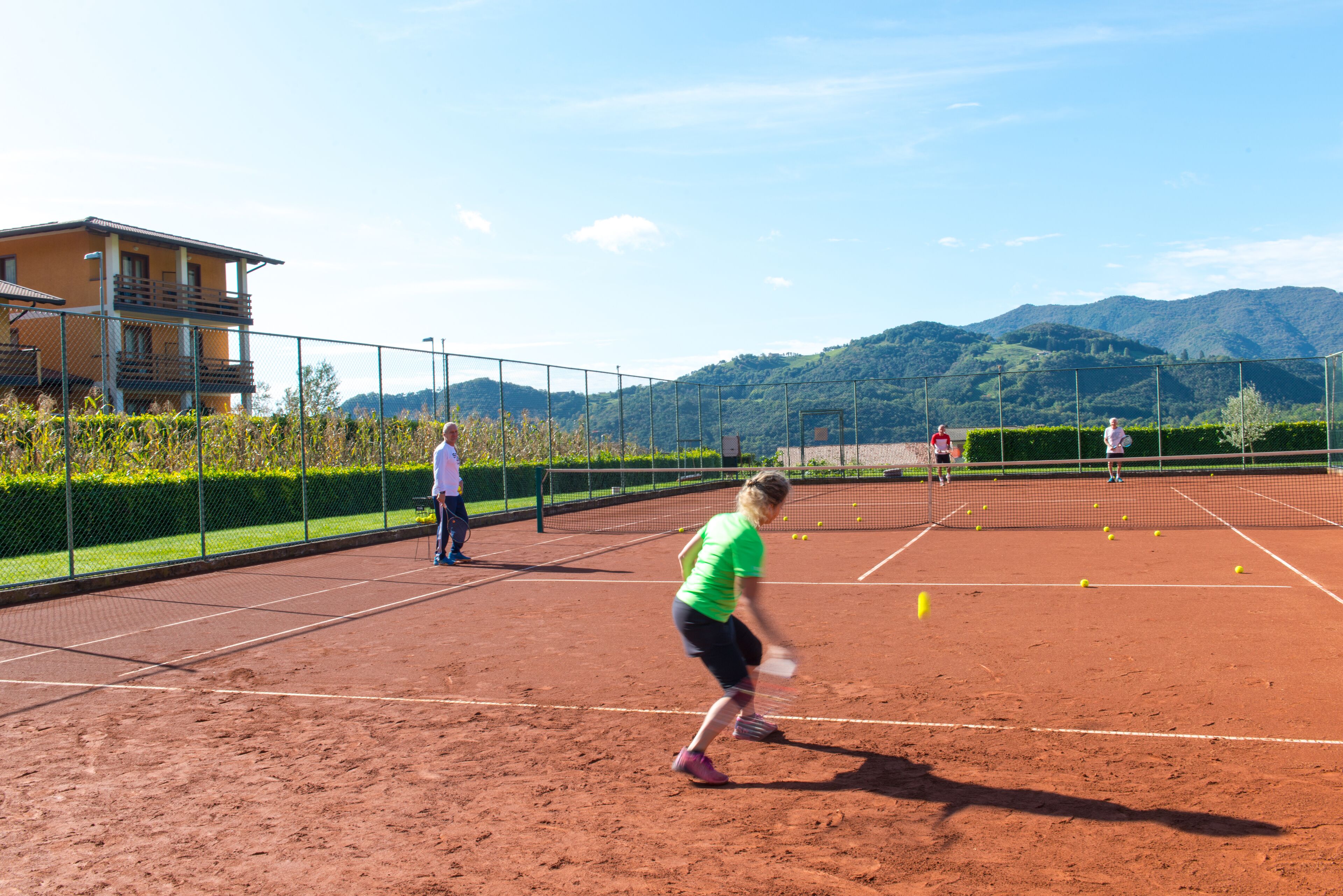En tennisbana med ett grusunderlag, omgiven av ett staket, och en byggnad med en balkong i bakgrunden.
