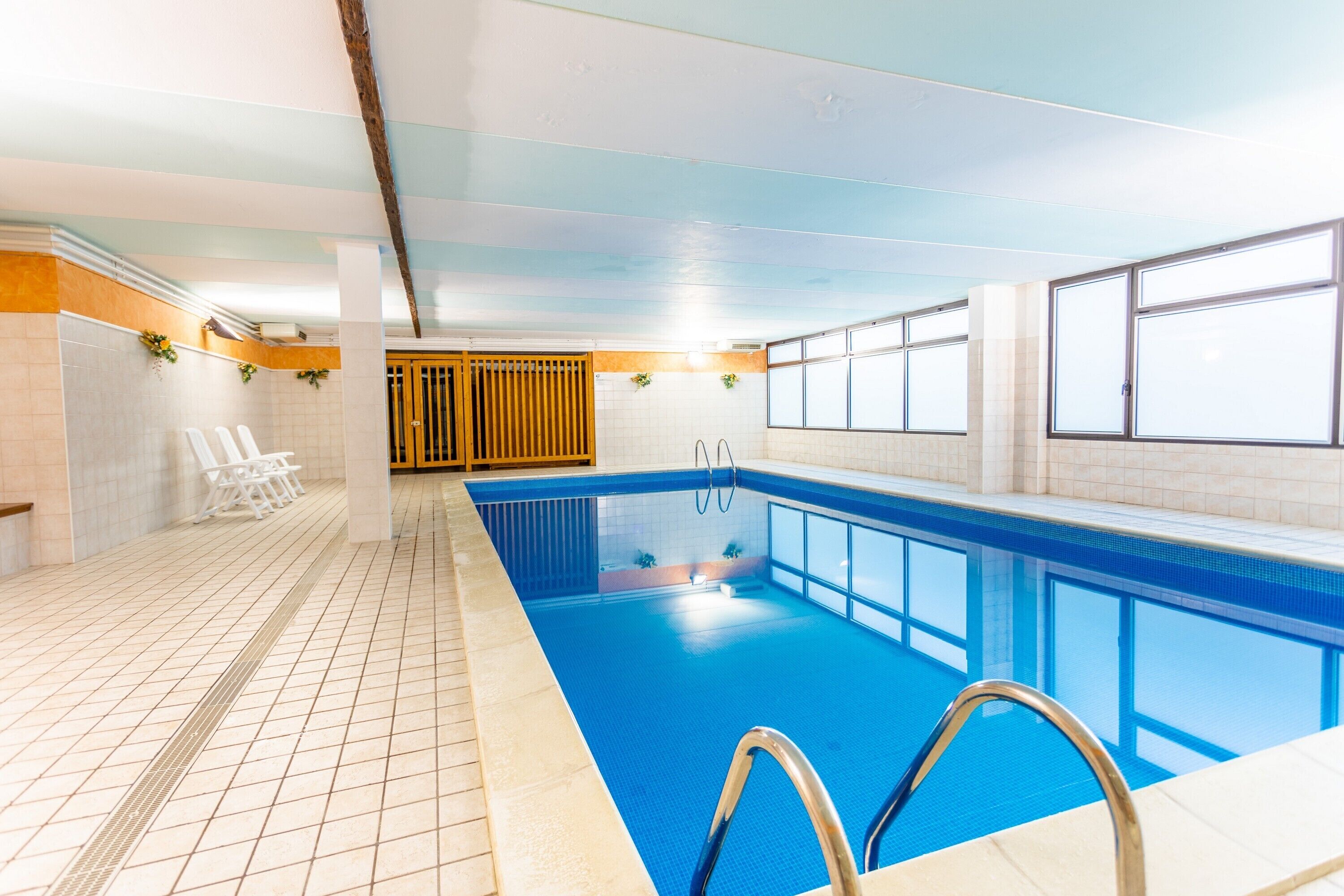An indoor swimming pool with clear blue water, tiled walls, and a tiled floor. There are lounge chairs and a wooden door in the background.