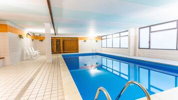 An indoor swimming pool with clear blue water, tiled walls, and a tiled floor. There are lounge chairs and a wooden door in the background.