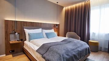 A modern hotel room with a wooden headboard, a large bed with white and blue bedding, a grey throw blanket, a bedside table with a lamp, and a round side table.
