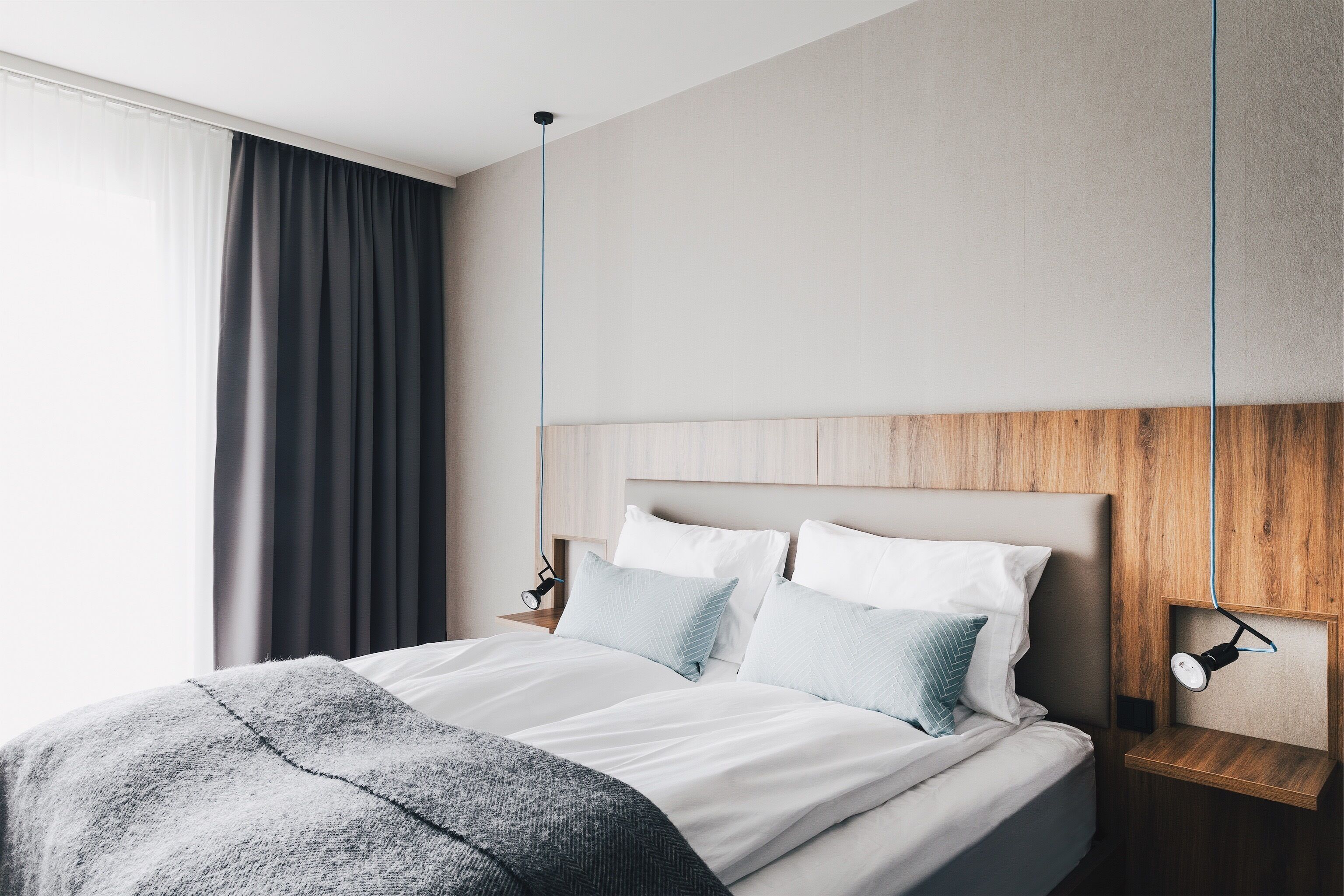 Ein modernes Hotelzimmer mit einem Holzkopfteil, einem Nachttisch mit Lampe und einem großen Fenster mit Vorhängen.