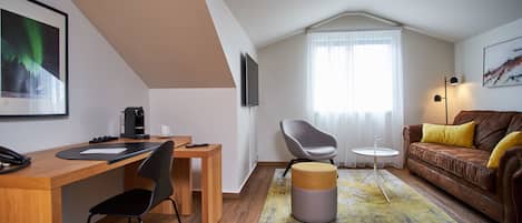 A modern living room with a wooden desk, a grey chair, a brown leather sofa with yellow cushions, a round ottoman, and a large window with sheer curtains.