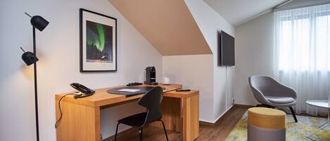 Una habitación moderna con un escritorio de madera, una silla de oficina negra y un otoman pequeño y redondo. Un cuadro enmarcado de la aurora boreal cuelga en la pared, y un televisor de pantalla plana está montado en la pared.