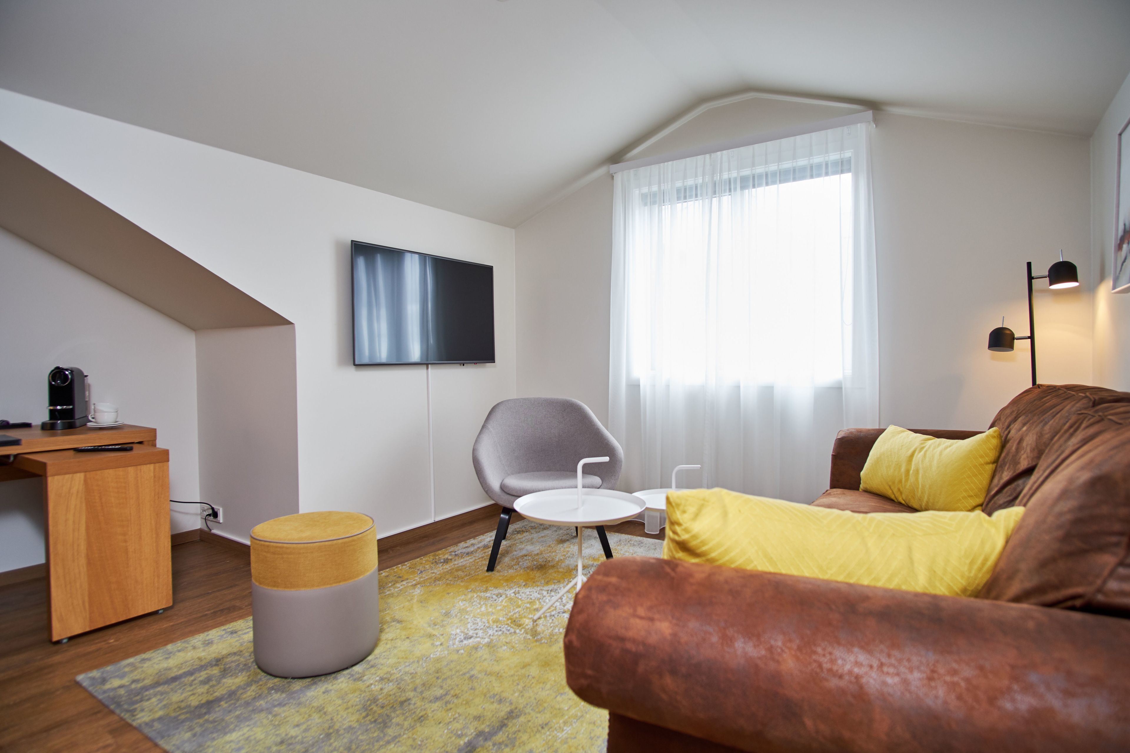 Un salon moderne avec un canapé marron, un fauteuil gris, un petit bureau en bois et un téléviseur à écran plat fixé au mur.