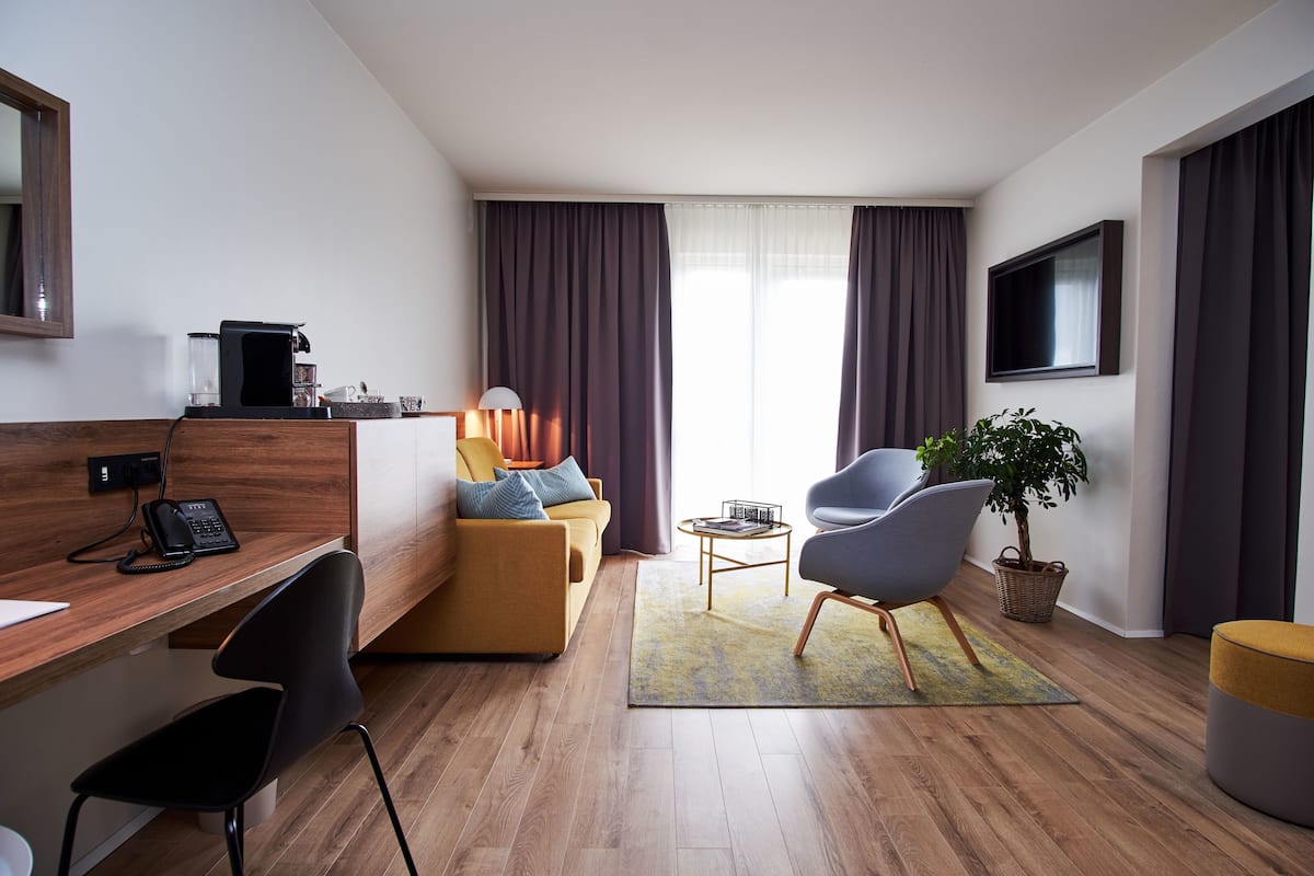 A modern hotel room with a wooden desk, a sofa, a chair, a small table, and a TV mounted on the wall.