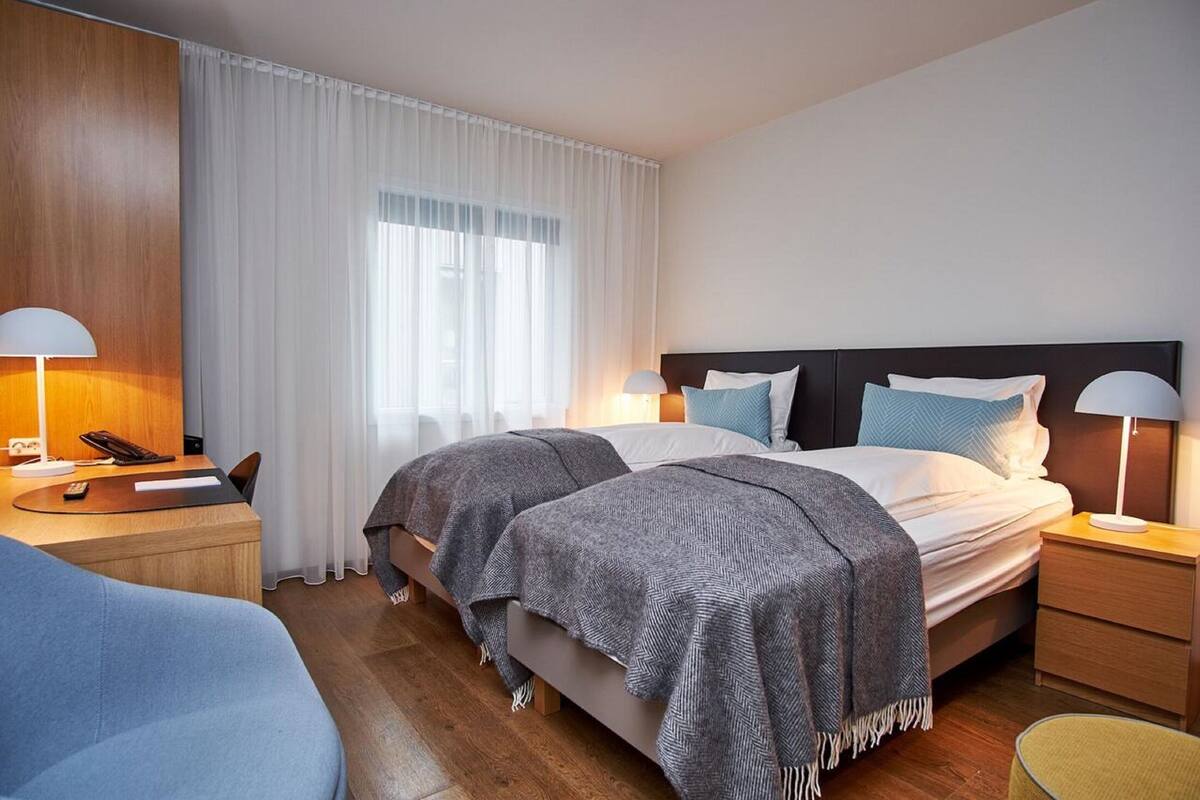 A hotel room with a double bed, two bedside tables with lamps, a desk with a chair, and a large window with sheer curtains.