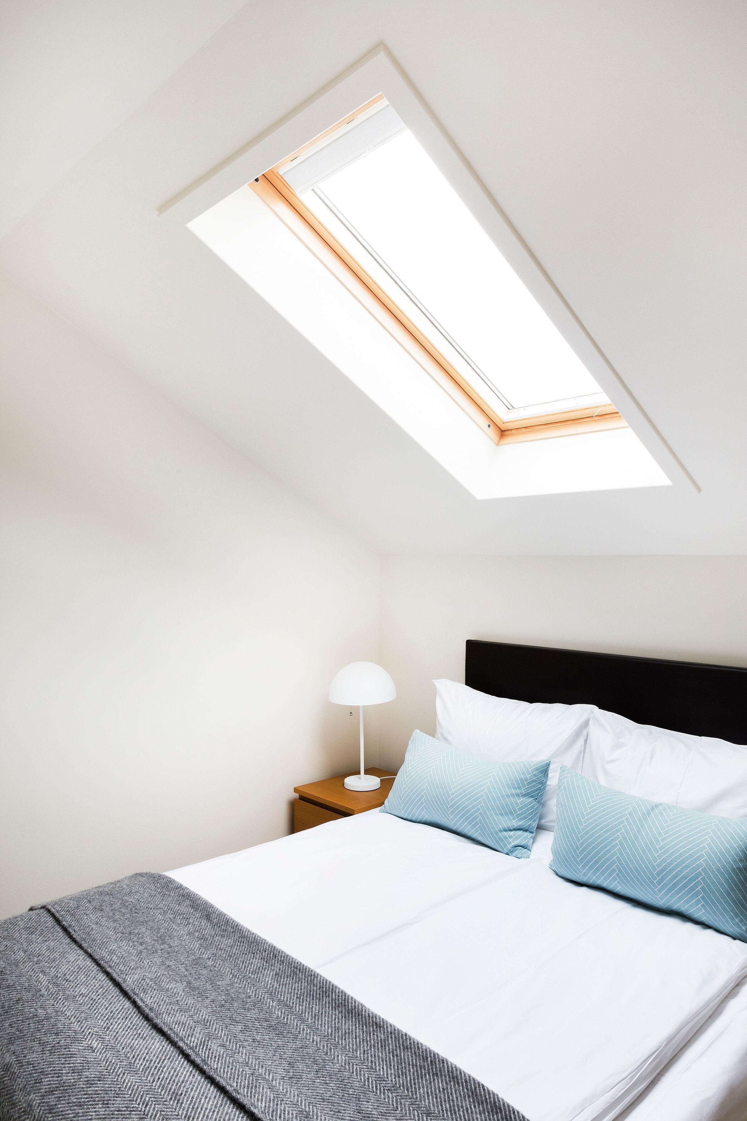 Un dormitorio con tragaluz, una cama con ropa de cama blanca y azul, una mesita de noche con lámpara y una manta de lana de punto color gris.
