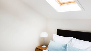 Un dormitorio con tragaluz, una cama con ropa de cama blanca y azul, una mesita de noche con lámpara y una manta de lana de punto color gris.