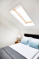 Un dormitorio con tragaluz, una cama con ropa de cama blanca y azul, una mesita de noche con lámpara y una manta de lana de punto color gris.