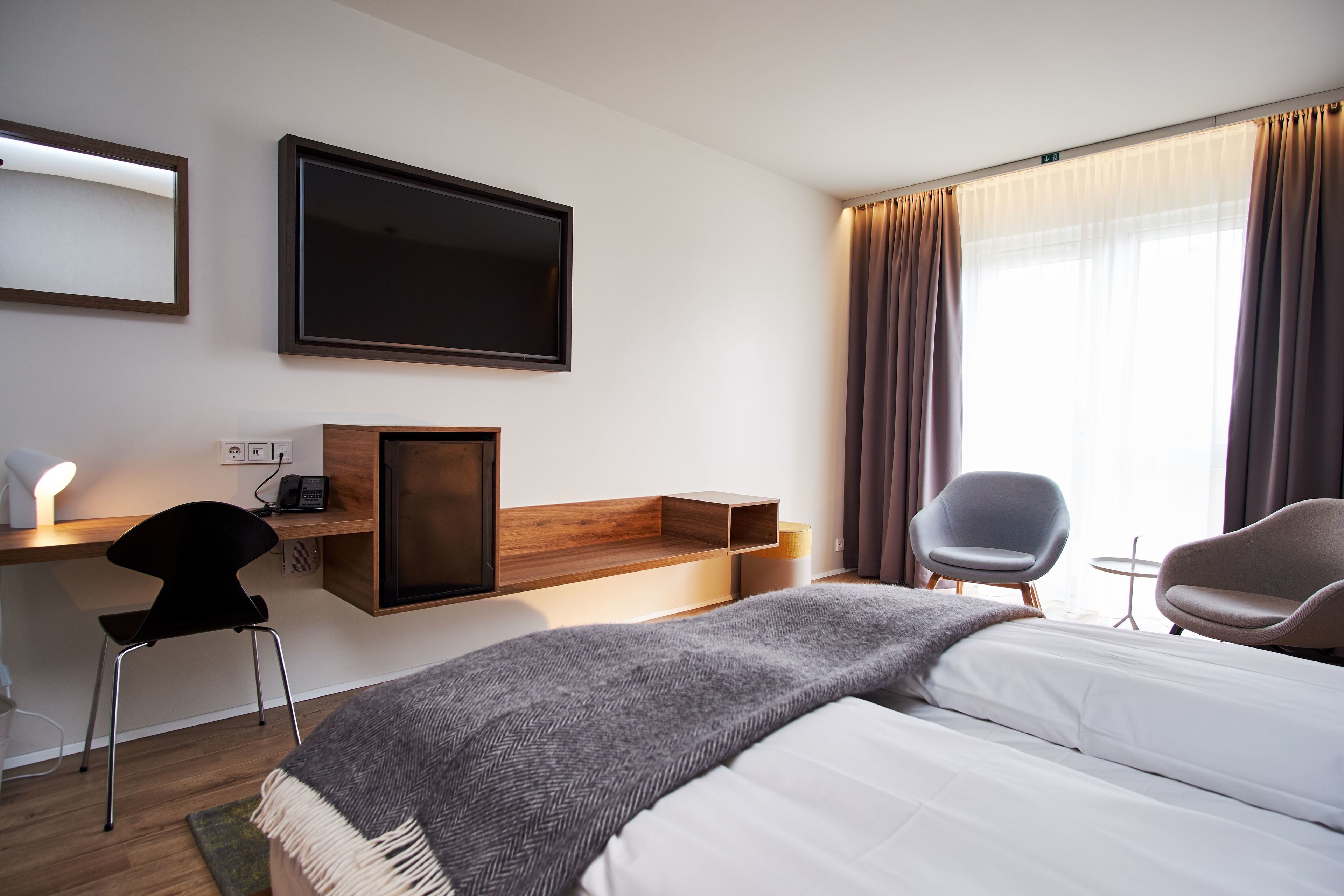 A modern hotel room with a large bed, a desk with a chair, a flat-screen TV, and a window with curtains.