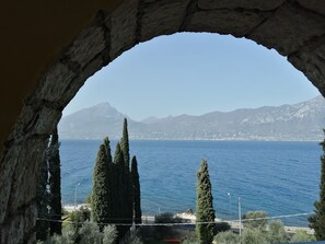 A view through a stone archway overlooking a coastal landscape with tall cypress trees and a distant mountain range.