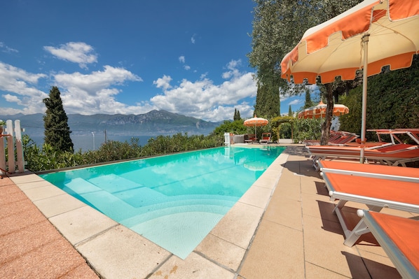 A swimming pool with lounge chairs and umbrellas, surrounded by greenery and a view of mountains.