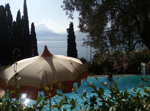 Área de piscina com um guarda-sol grande, cercada por vegetação e com vista para um corpo d'água com montanhas ao longe.