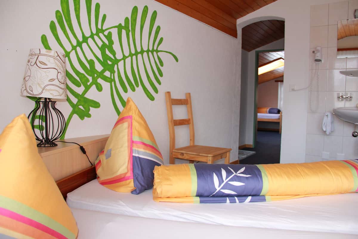 A hotel room with a bed, pillows, a lamp, a wooden chair, and a bathroom visible through an archway.