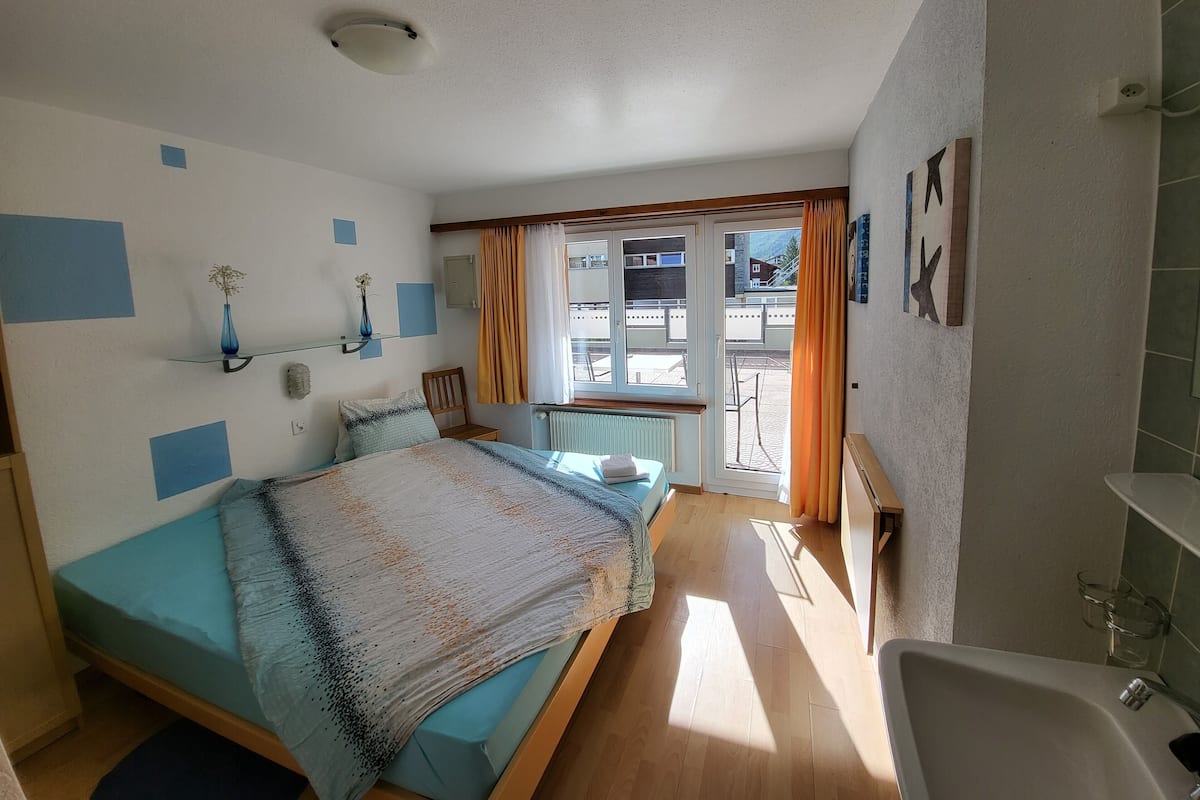 A bedroom with a bed, a chair, a small table, a radiator, a window with curtains, and a view of the outside.