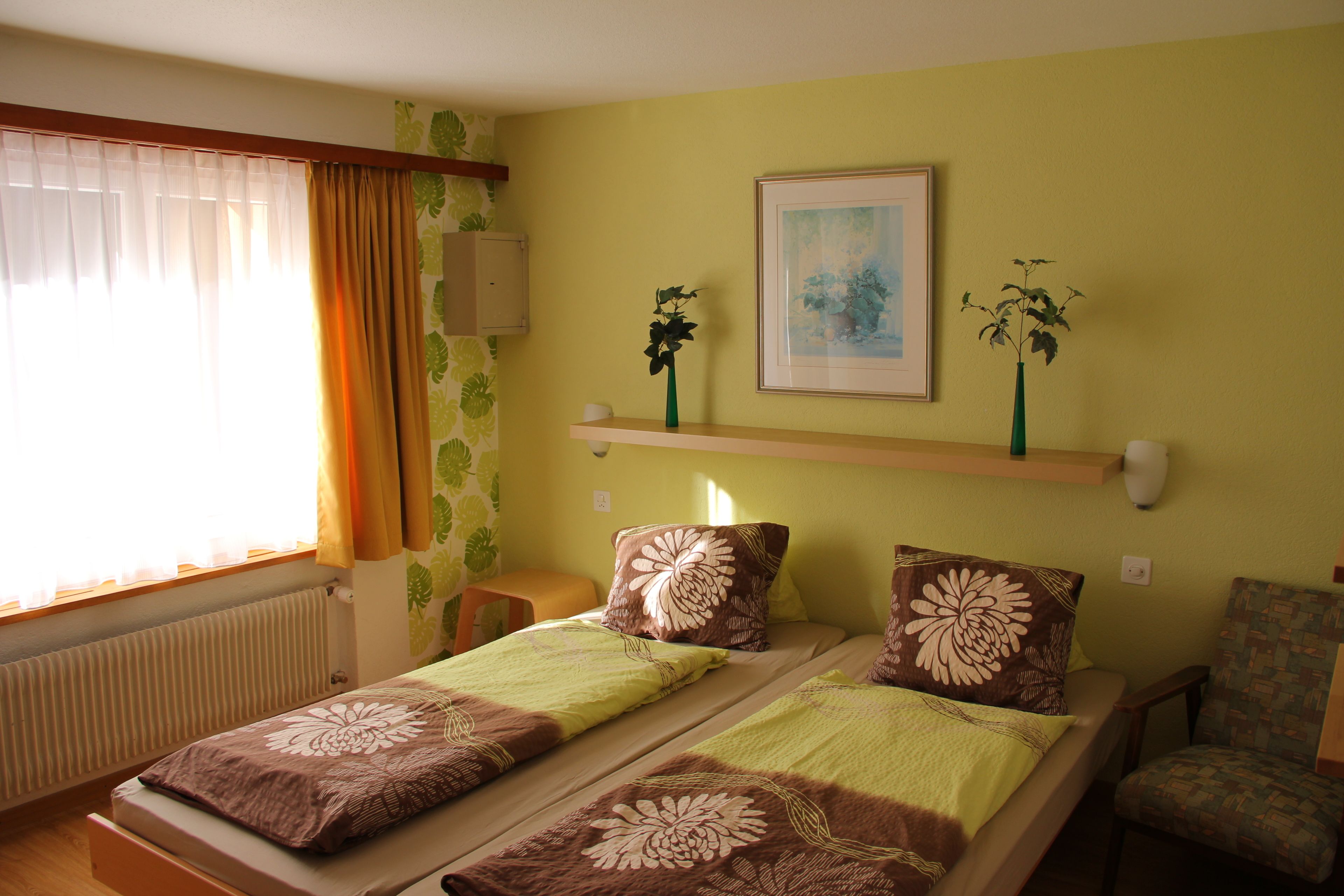 A hotel room with two beds, a chair, a shelf with decorative items, and a window with curtains.