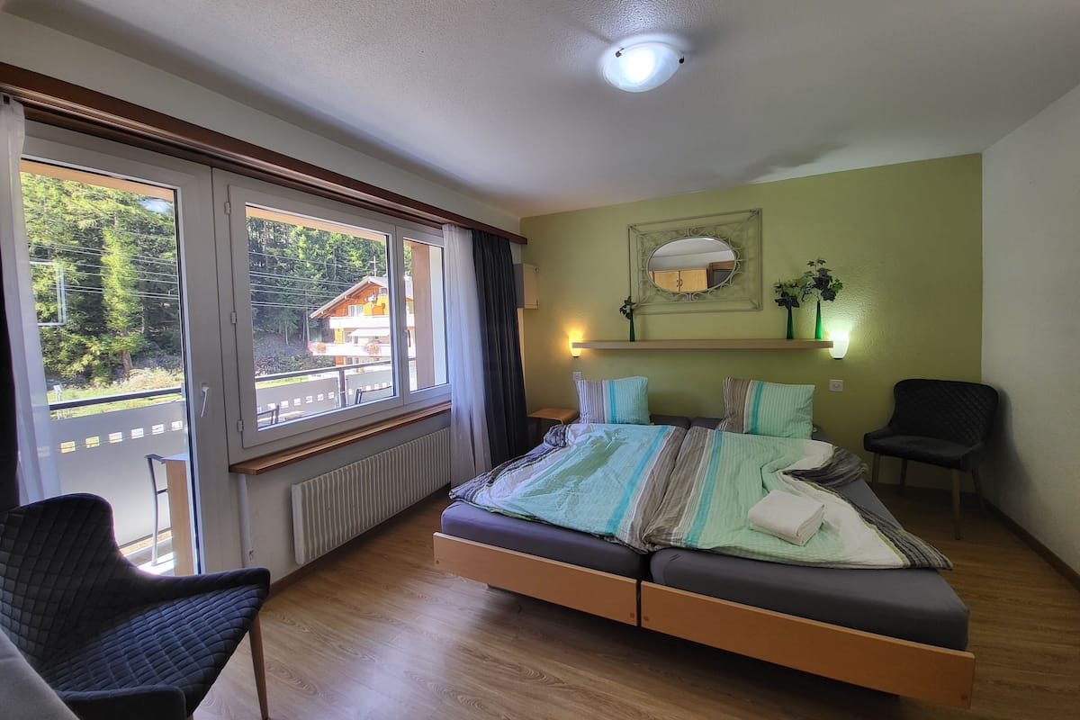 A bedroom with a bed, two chairs, a window with curtains, a radiator, a ceiling light, and a mirror on the wall.
