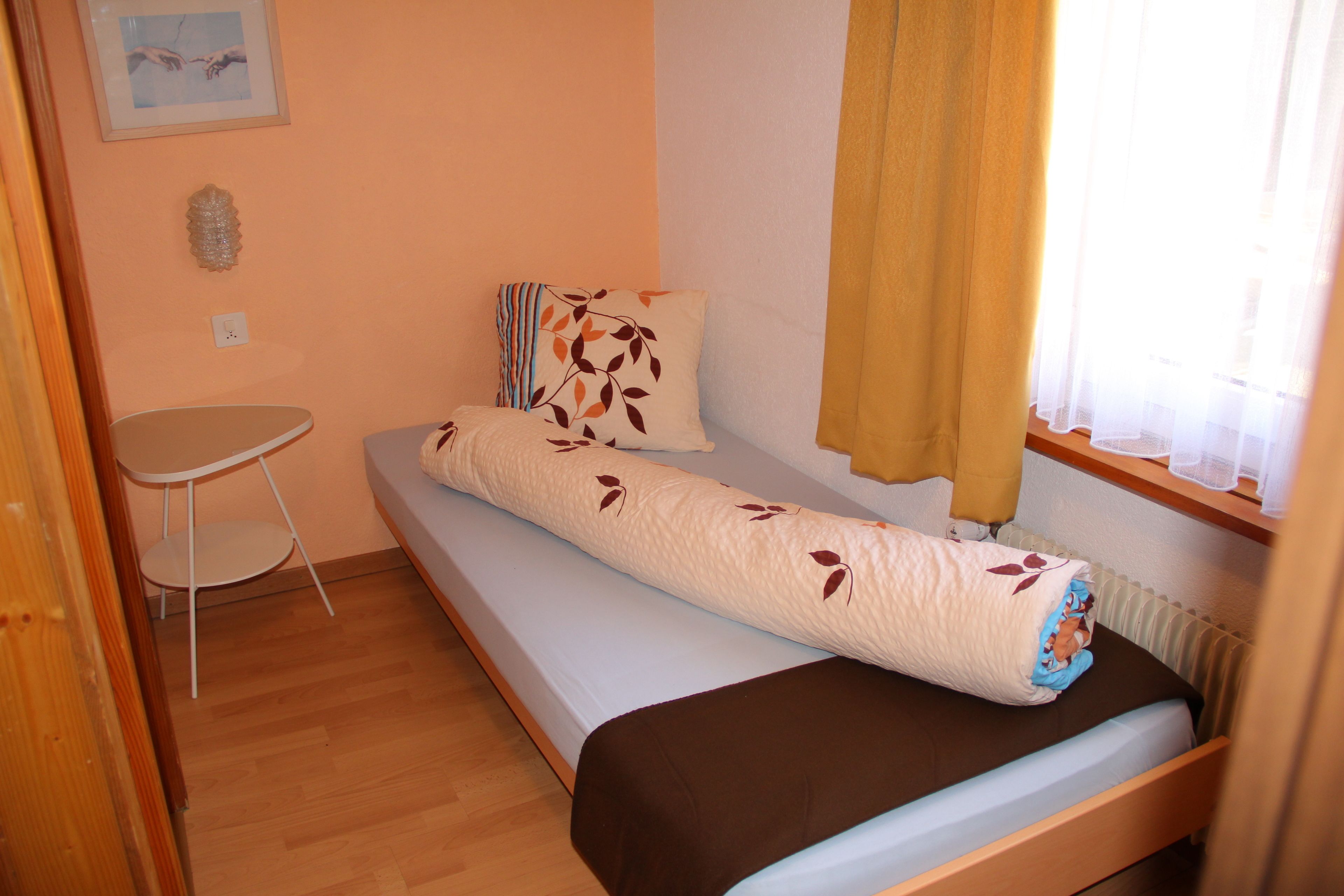 A small, single-bed room with a wooden floor, a small table, a radiator, and a window with curtains.