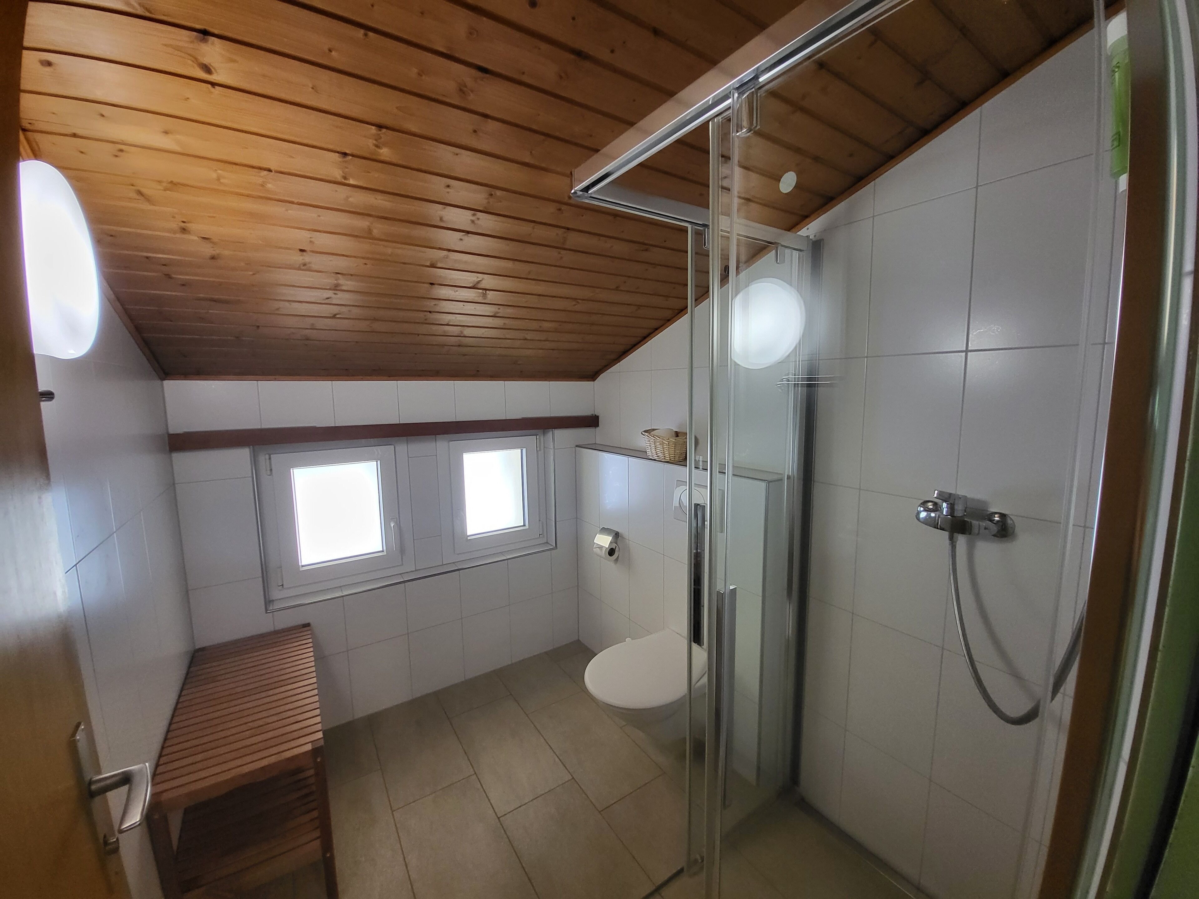 Egy fürdőszoba, melyben fa borítja a mennyezetet, üvegfalas zuhanyzó, WC és egy pad található.