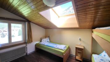 A small, cozy room with a sloped ceiling and a skylight, a single bed with a green mattress, a wooden nightstand, and a radiator.