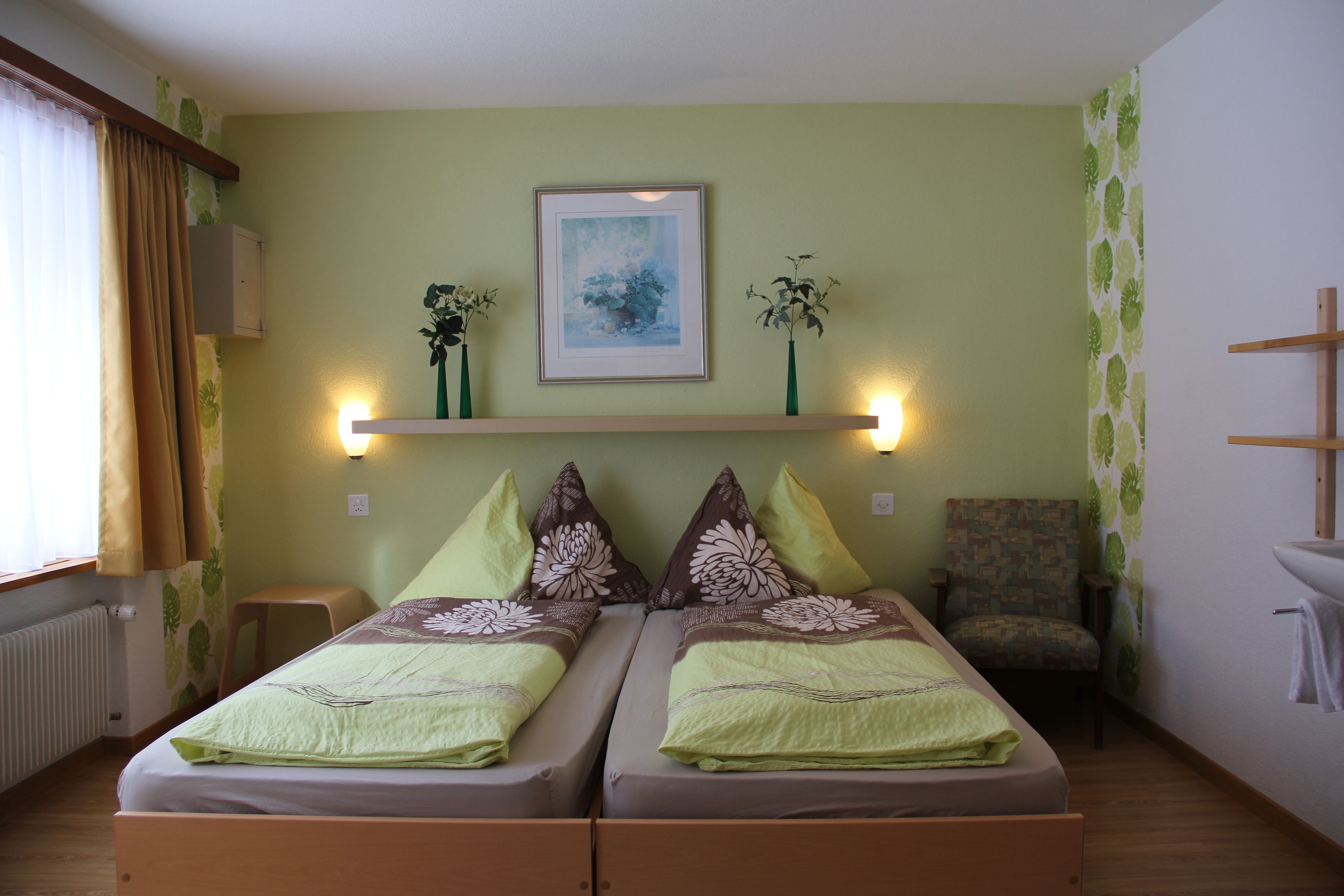 A twin room with two beds, a chair, a small table, a radiator, and a framed picture on the wall.