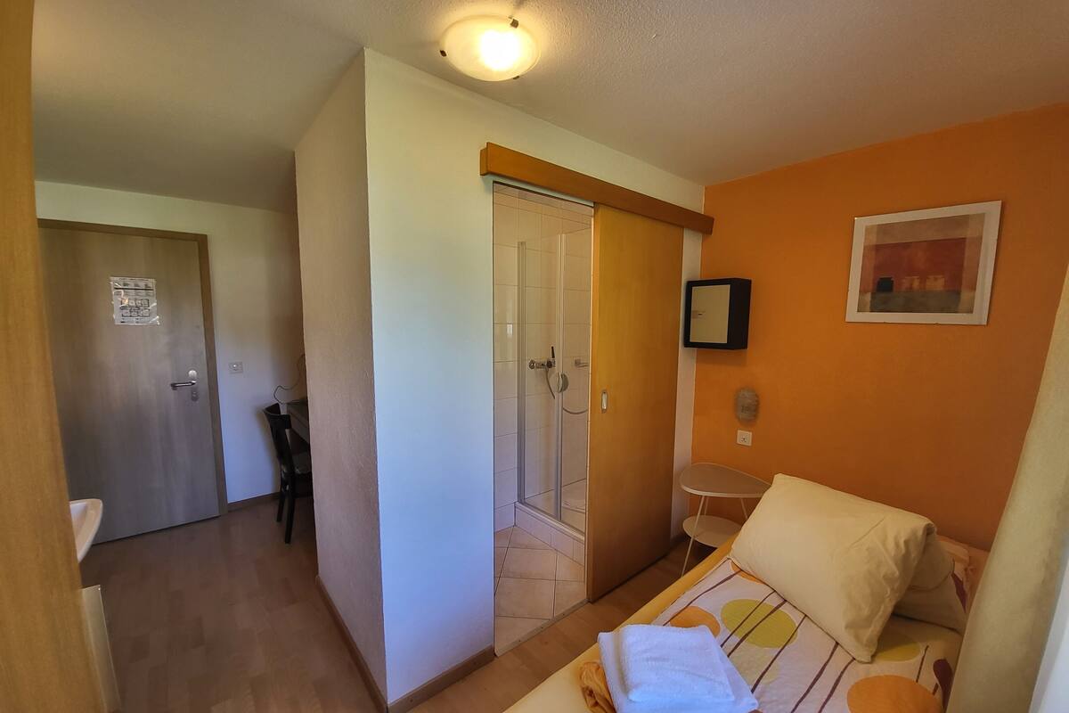 A hotel room with a bed, a chair, a small table, a shower area, and a door with a sign.