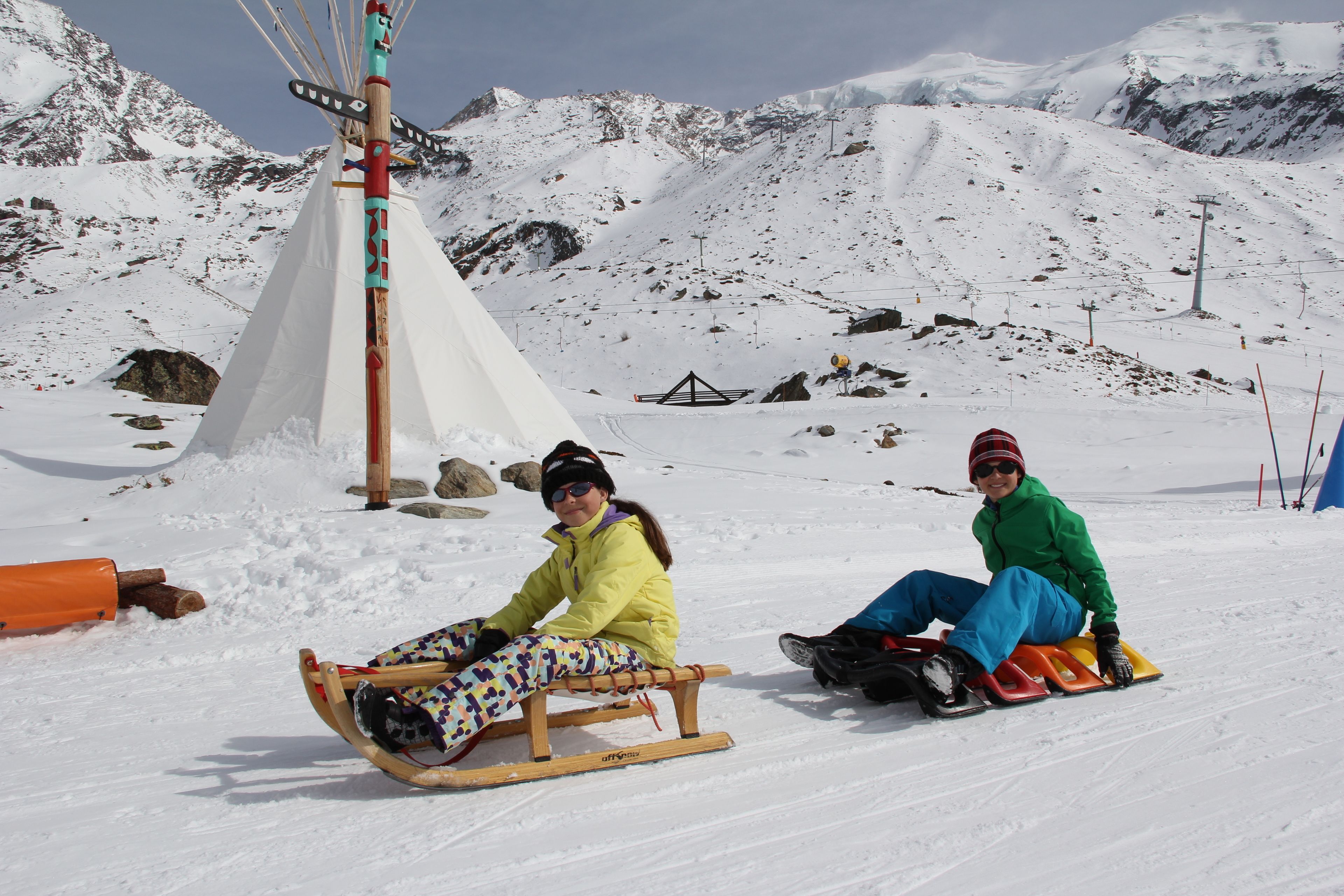 Two people sledding on a snowy slope with a mountain backdrop.