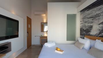 Una habitación de hotel moderna con un televisor de pantalla plana grande, una cama bien hecha con almohadas, una mesita de noche con un libro y un frutero, y un cuadro abstracto grande en la pared.