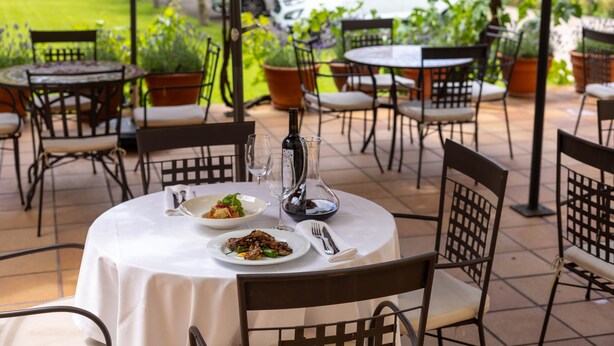 An outdoor dining area with round tables and chairs, a white tablecloth, and a bottle of wine.