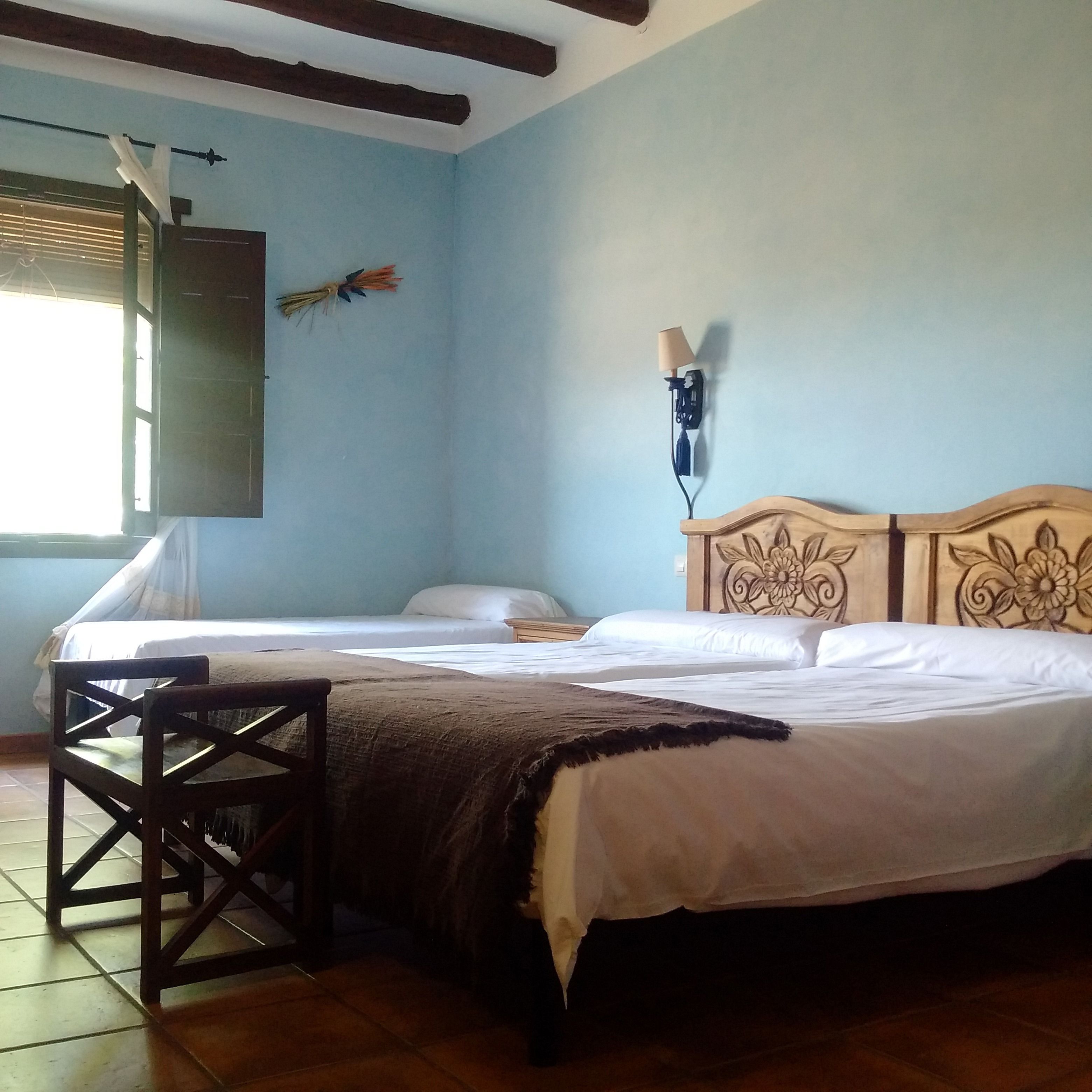 Une chambre avec deux lits, une tête de lit en bois, une table de chevet, une chaise, une lampe fixée au mur et une fenêtre avec des rideaux.