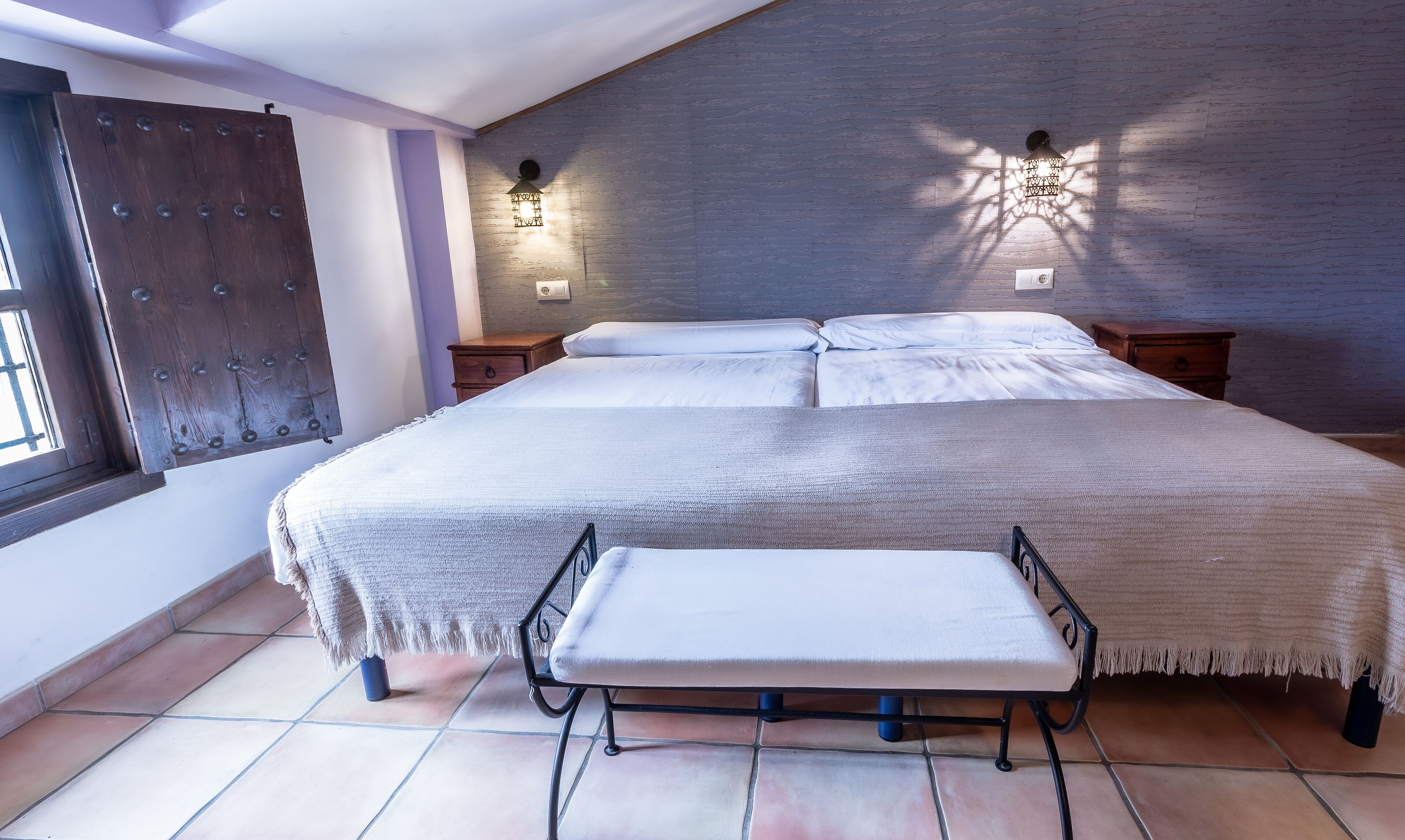 A bedroom with a large bed, a bench, two nightstands, a wall-mounted light fixture, and a window with a wooden door.