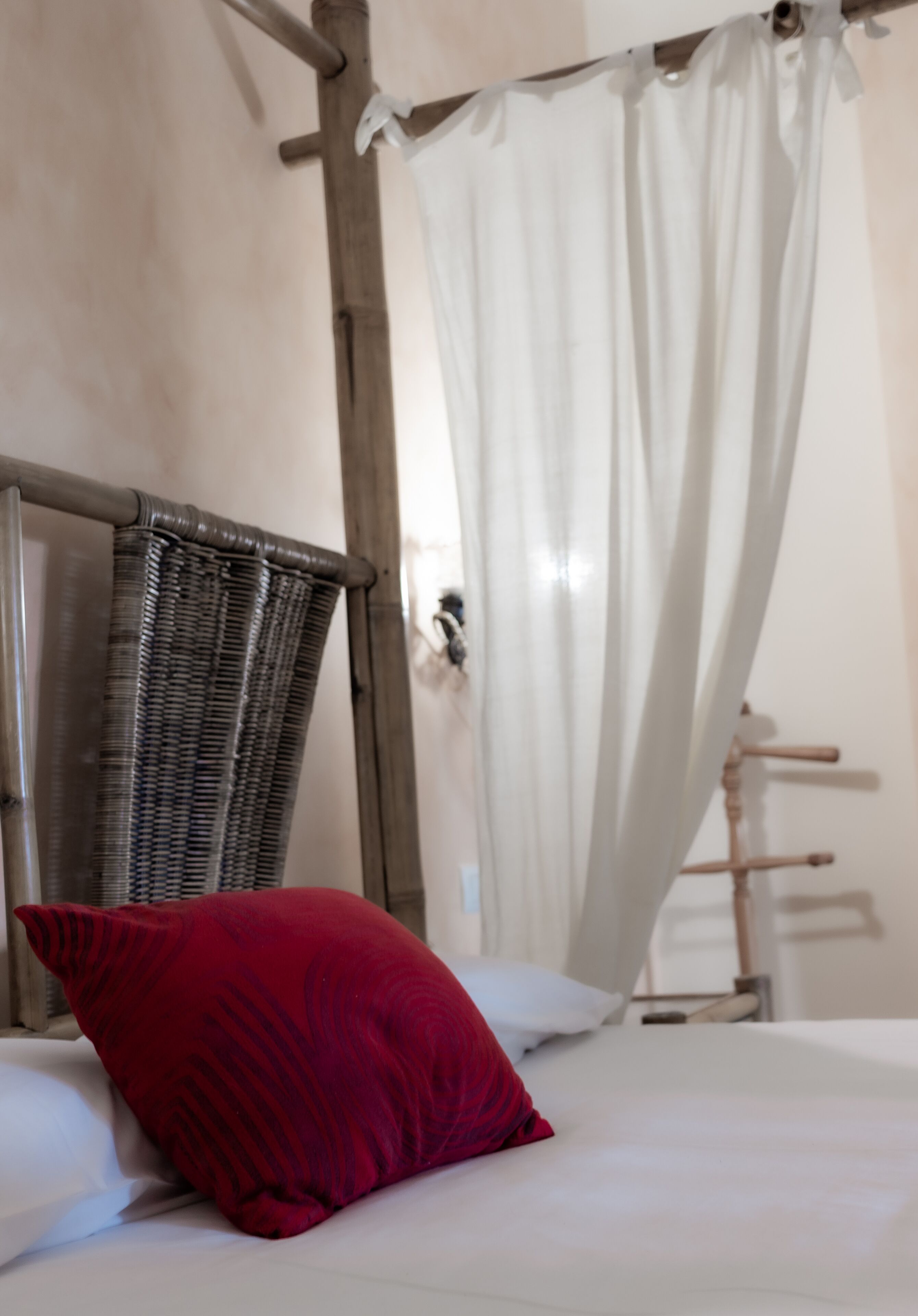 Una cama con sábanas blancas, un almohadón rojo y una silla de mimbre.